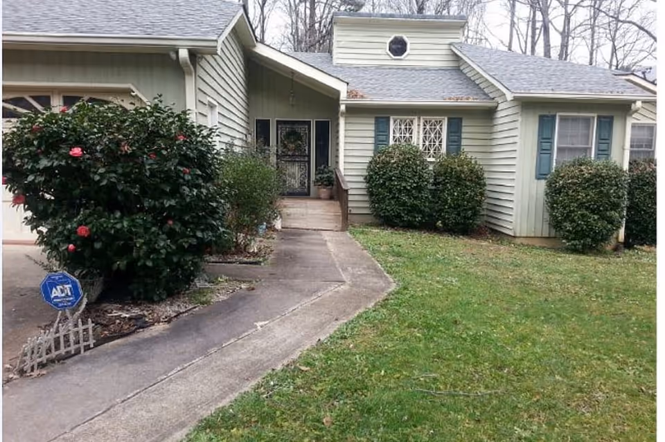 Front exterior view of a single-story house with light green siding, blue shutters, and a small porch with steps leading to a black front door. There are bushes and a flowering shrub near the walkway, and a green lawn in front. Trees without leaves are visible in the background.