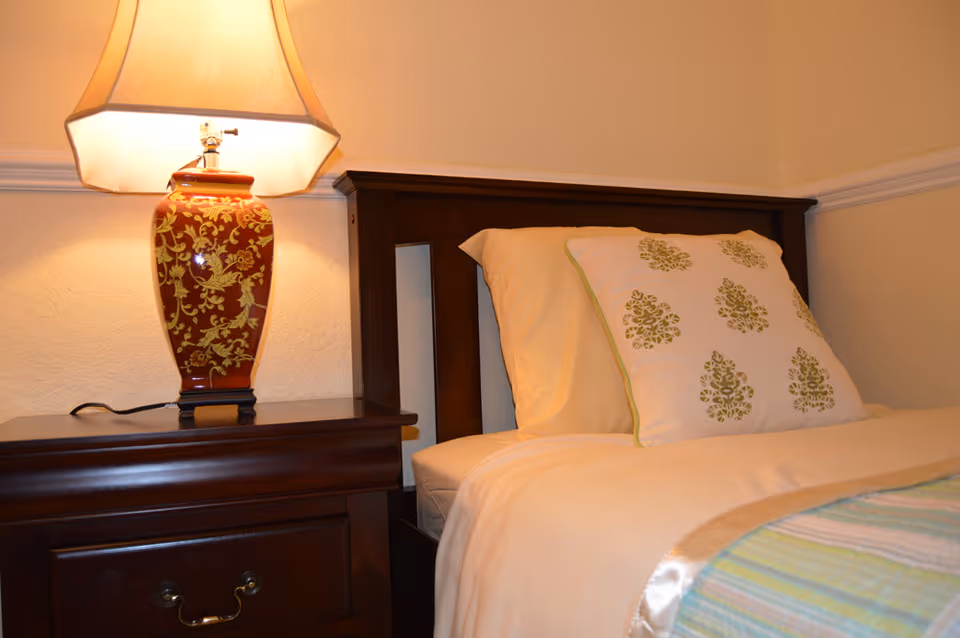 A close-up view of a neatly made bed with a wooden headboard, cream-colored sheets, and a decorative pillow with green floral patterns. Next to the bed is a dark wooden nightstand with a red and gold patterned lamp emitting warm light.