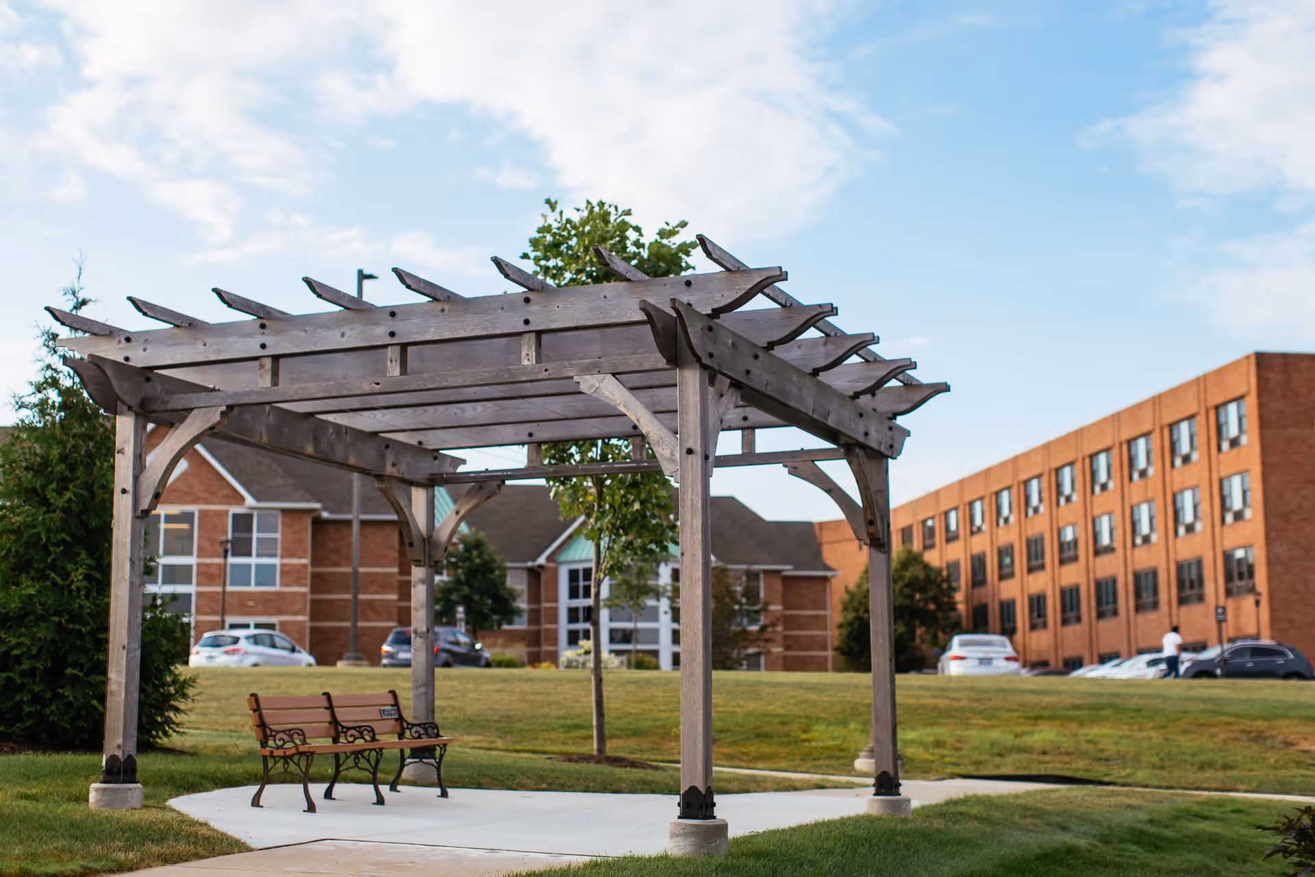 Wooden pergola with a bench on a grassy courtyard in front of brick senior living buildings under a partly cloudy sky.