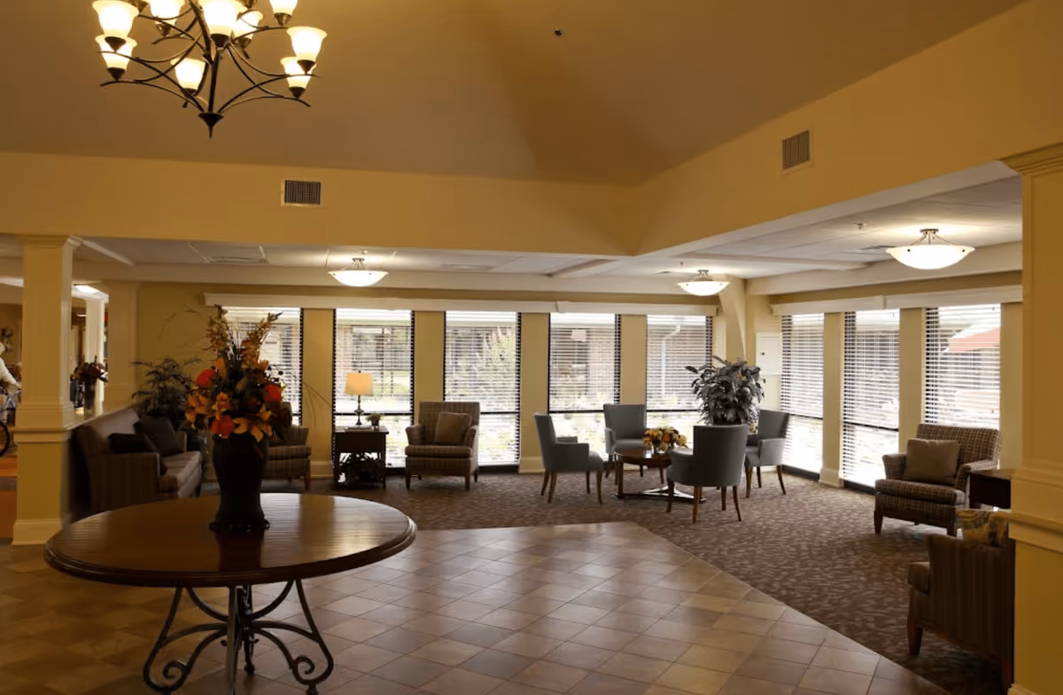A spacious, well-lit living room area with multiple seating arrangements including armchairs and sofas. Large windows with blinds allow natural light to fill the room. A round wooden table with a floral arrangement is in the foreground, and ceiling lights and a chandelier provide additional lighting.