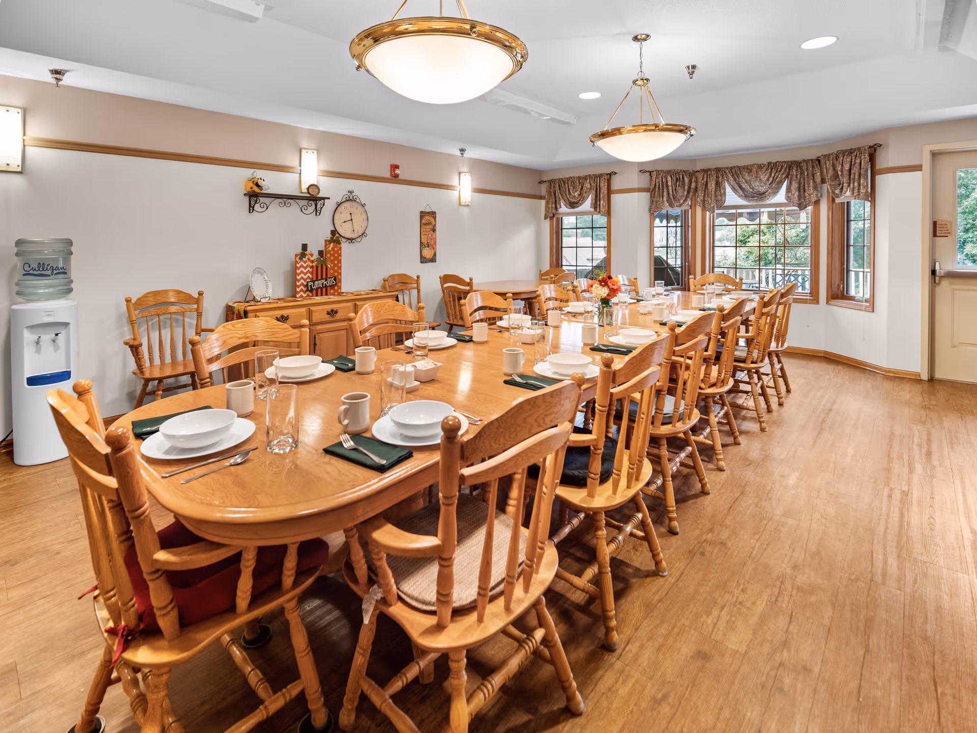 A long wooden dining table set with white plates, bowls, mugs, glasses, and silverware with green napkins in a bright dining room. The room has wooden chairs around the table, large windows with brown curtains, wooden flooring, and two ceiling light fixtures. A water cooler is visible in the corner, and a wooden sideboard with decorative items is against the wall.