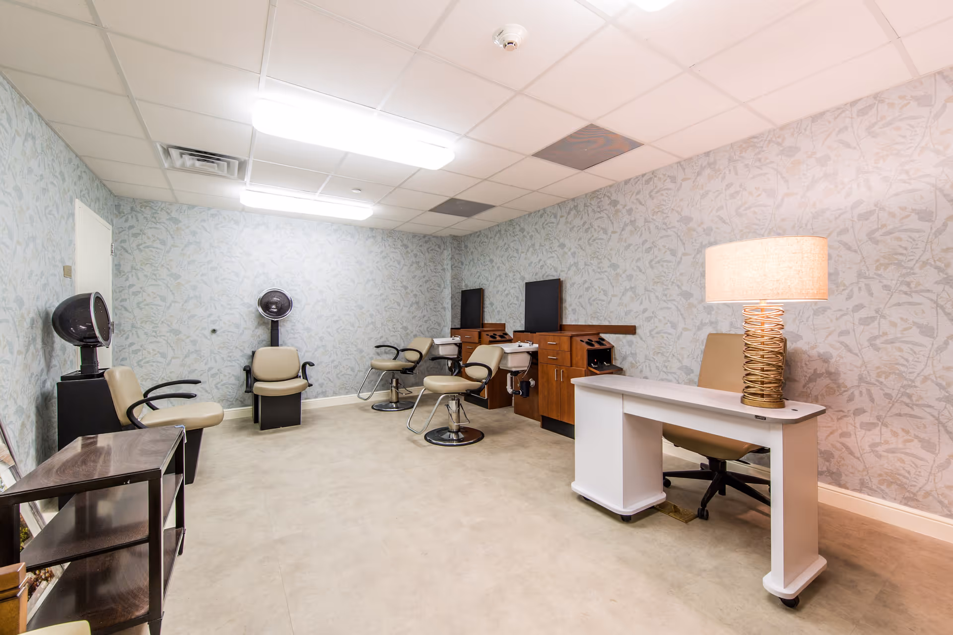 A small salon room with three salon chairs, two hair dryers, a white desk with a lamp, and a beige chair. The walls have a light floral pattern wallpaper and the floor is covered with beige carpet.