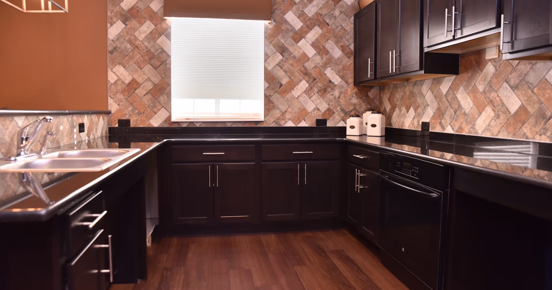 Modern kitchen with dark wood cabinets, black granite countertops, a stainless steel sink, and a window with a white blind. The backsplash features a herringbone pattern of beige and brown tiles, and the floor is a dark wood finish.