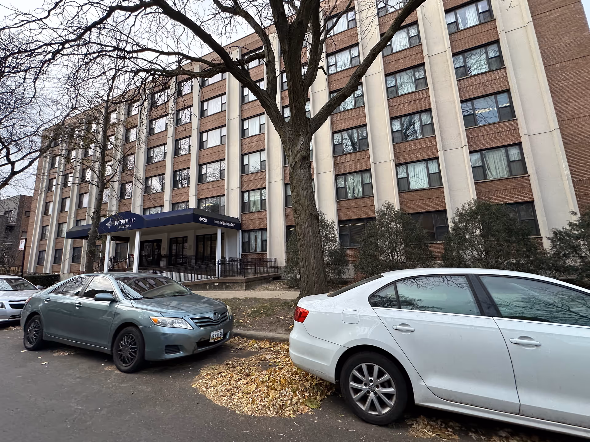 Exterior of a multi-story brick-and-concrete senior living building with a covered entrance, parked cars, and a large tree in front.
