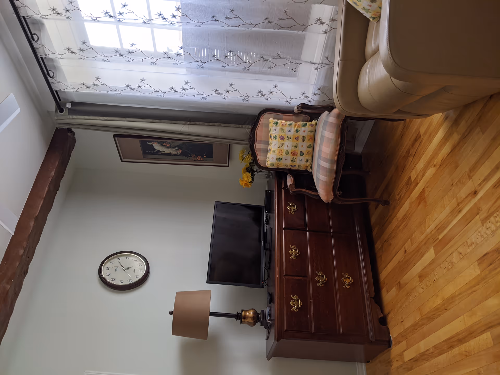 A cozy living room corner with a wooden dresser holding a flat-screen TV and a table lamp. Next to the dresser is an upholstered armchair with a floral cushion. A beige leather sofa is partially visible. The room has wooden flooring, a wall clock, a framed picture, and a window with sheer curtains featuring a floral pattern.