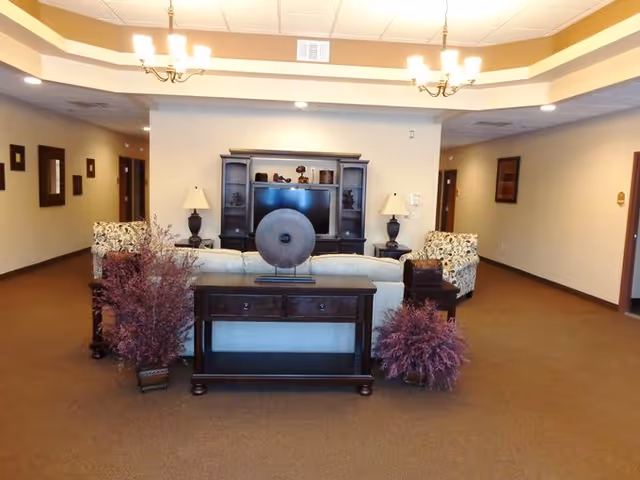 A spacious living room area in an assisted living facility featuring a beige sofa with patterned armchairs on either side. In front of the sofa is a dark wooden console table with decorative items and two potted plants with purple foliage on the floor. The back wall has a large entertainment center with a flat-screen TV and two table lamps on either side. The ceiling has recessed lighting and two chandeliers. The room has beige walls and carpeted flooring with doorways leading to other rooms.