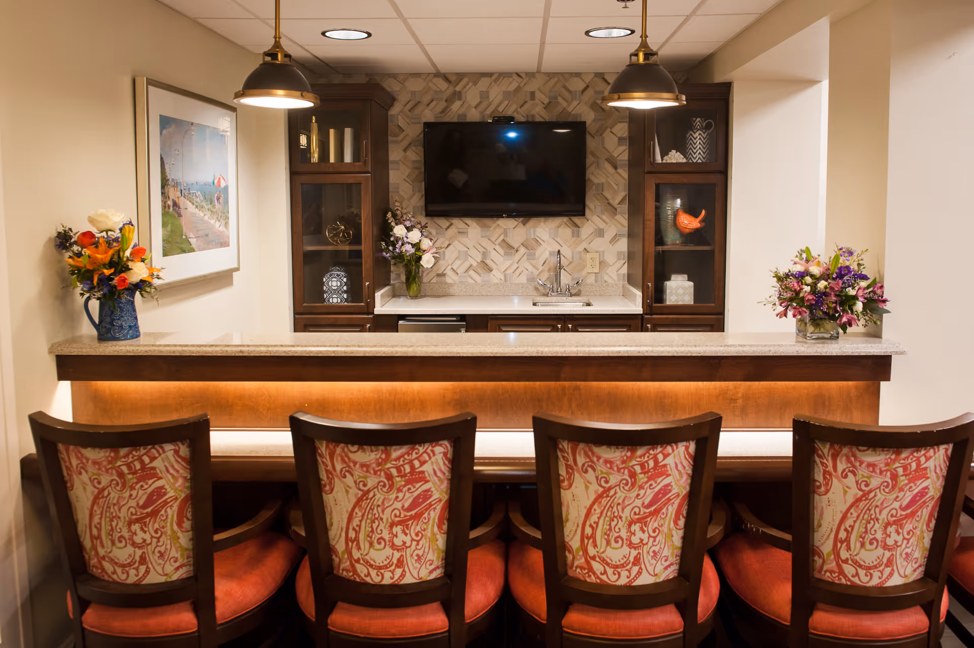 Cozy bar-style kitchenette with five upholstered chairs facing a counter, wall-mounted TV, cabinets, a sink and floral arrangements.