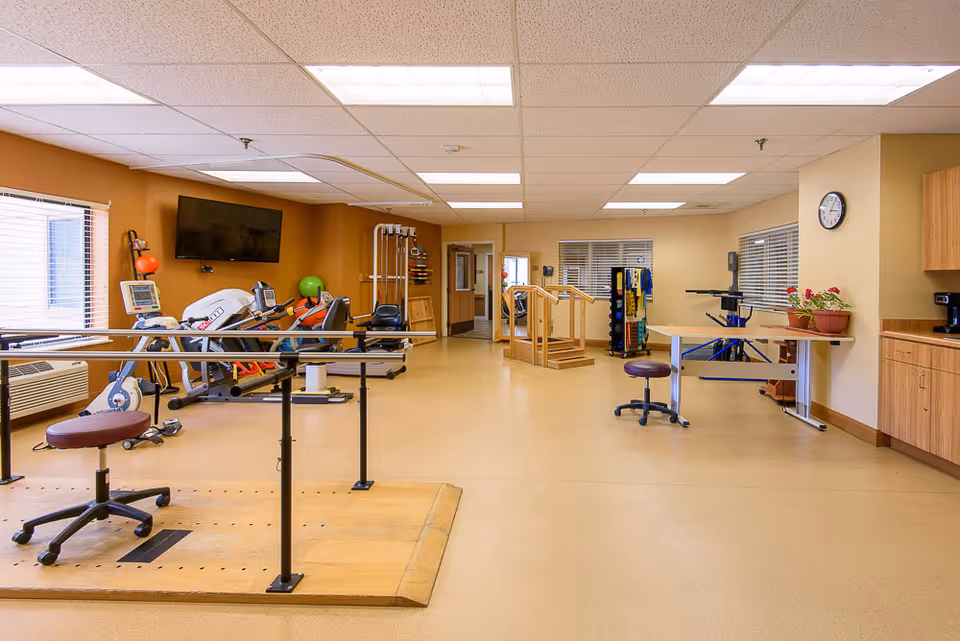 A spacious physical therapy room with exercise equipment, parallel bars, therapy stairs, and seating in a senior care facility.