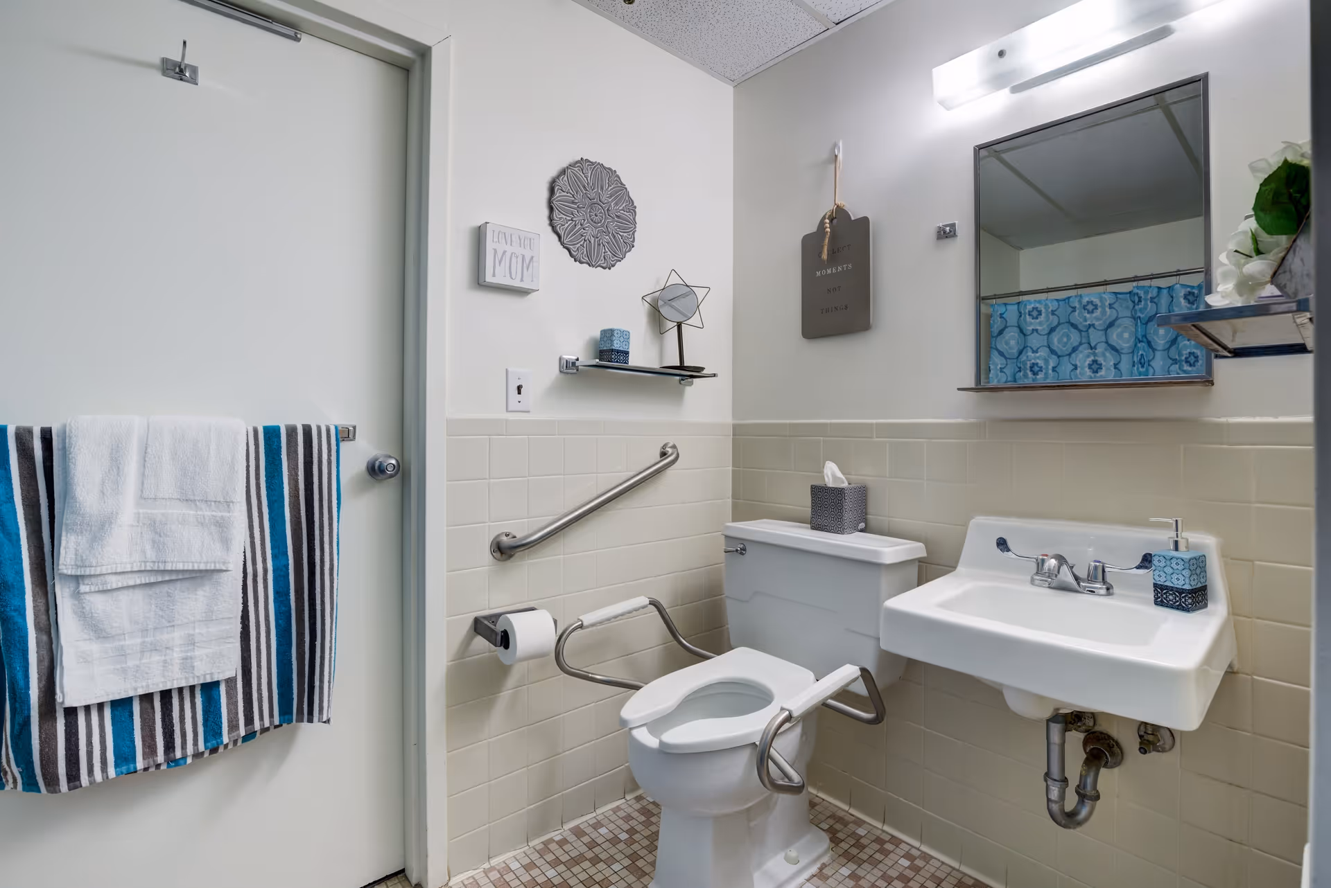 A clean, accessible bathroom with a white toilet equipped with safety rails, a white sink with a soap dispenser, a mirror above the sink, and a towel rack on the door holding a white towel and a striped towel. The walls are tiled halfway up and decorated with small wall art pieces.