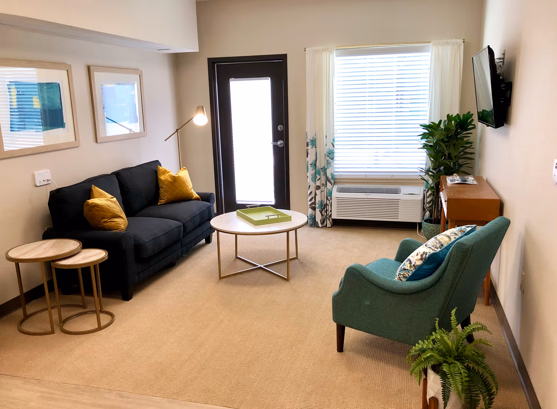 A cozy living room with a dark blue sofa adorned with yellow pillows, a green armchair with patterned cushions, a round coffee table with a green tray, two nested side tables, a floor lamp, a wall-mounted TV, a wooden console table, and two framed artworks on the wall. There is a window with white blinds and floral curtains, a door with a frosted glass panel, and two potted plants on the floor.
