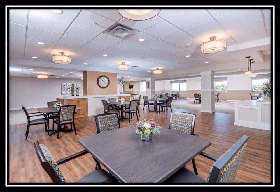 A spacious and well-lit dining area with multiple tables and chairs arranged neatly. The room features wood flooring, modern ceiling lights, a large wall clock, and decorative plants and flowers on the tables. Large windows allow natural light to brighten the space.