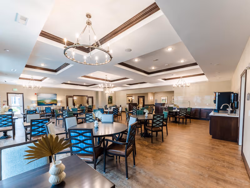 Spacious dining room in a senior living facility with multiple round tables and chairs arranged neatly. The room features wooden flooring, modern chandeliers hanging from a coffered ceiling, and decorative vases on the tables. There is a counter with a sink and coffee machine along one wall, and artwork and mirrors adorn the walls.