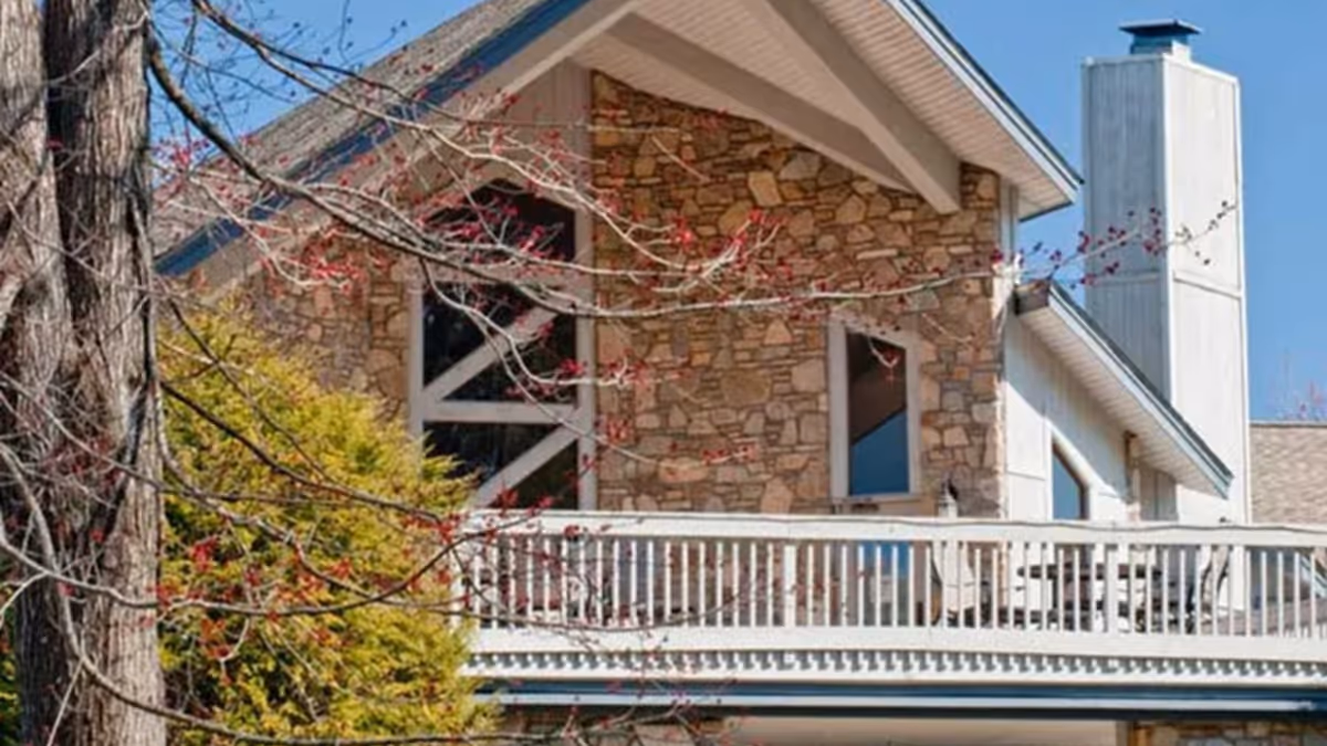 Exterior view of a senior living facility building with stone walls, large windows, a white balcony railing, and a chimney. Trees with budding leaves are visible in the foreground under a clear blue sky.