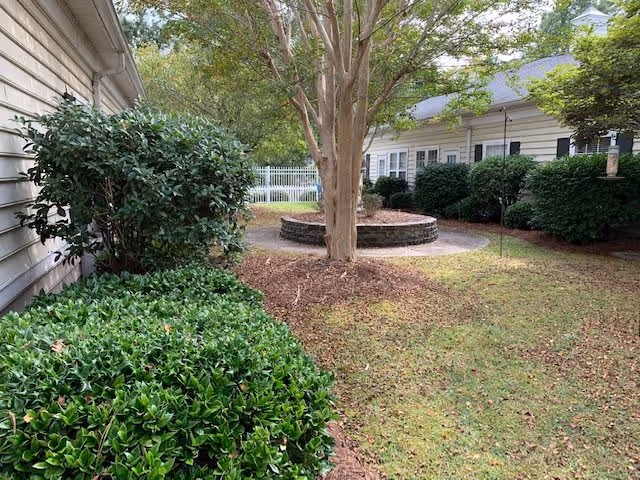 Outdoor garden area with green bushes and a tree in the center surrounded by a circular stone bench. White buildings with black shutters and a white fence are visible in the background.