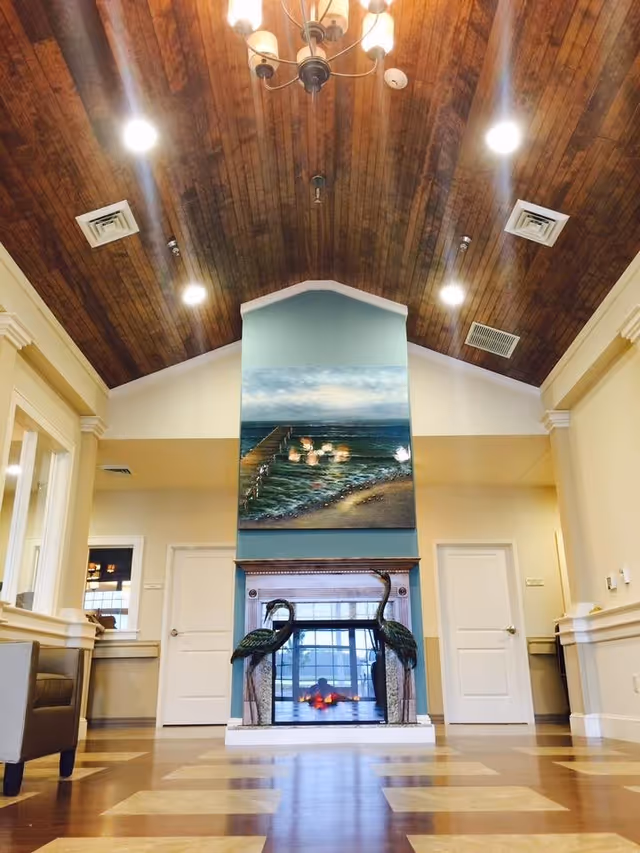 Interior view of a room with a high wooden ceiling and recessed lighting. The room features a central fireplace with decorative metal bird sculptures on either side and a painting of a pier over water above it. There are two white doors on either side of the fireplace and beige walls with white trim. A brown armchair is partially visible on the left side.