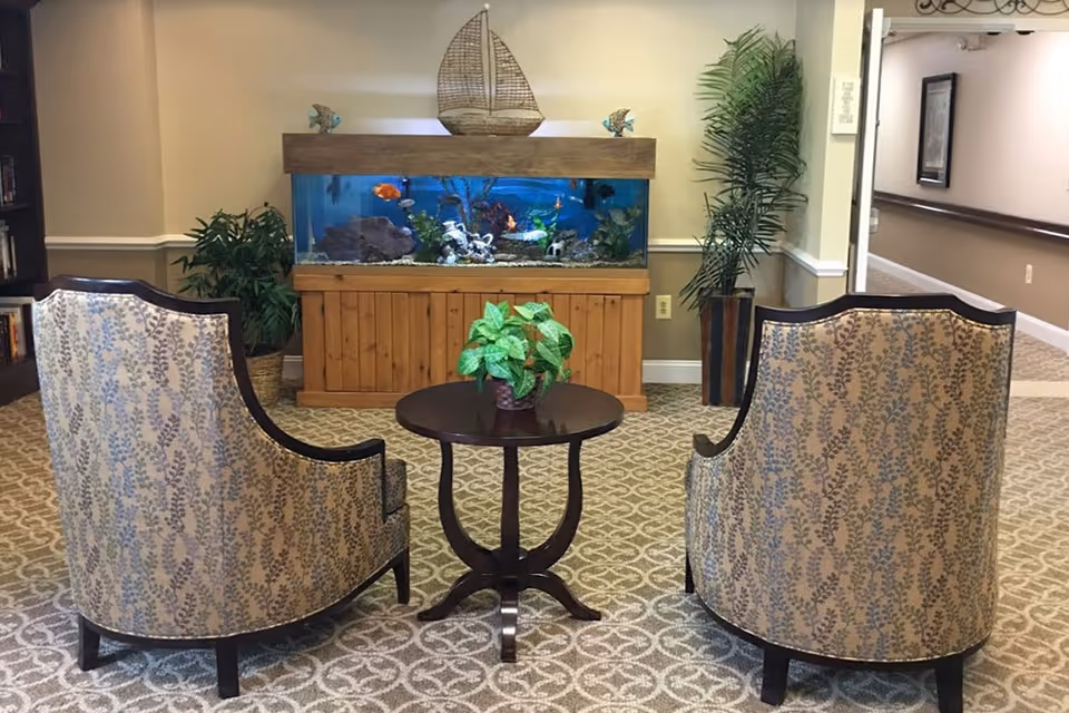 A cozy seating area in a senior living facility with two patterned armchairs facing a round wooden table with a potted plant. Behind the table is a large fish tank with colorful fish, decorative plants, and a wooden sailboat ornament on top. The room has beige walls, patterned carpet, and potted plants on either side of the fish tank. A hallway is visible to the right.