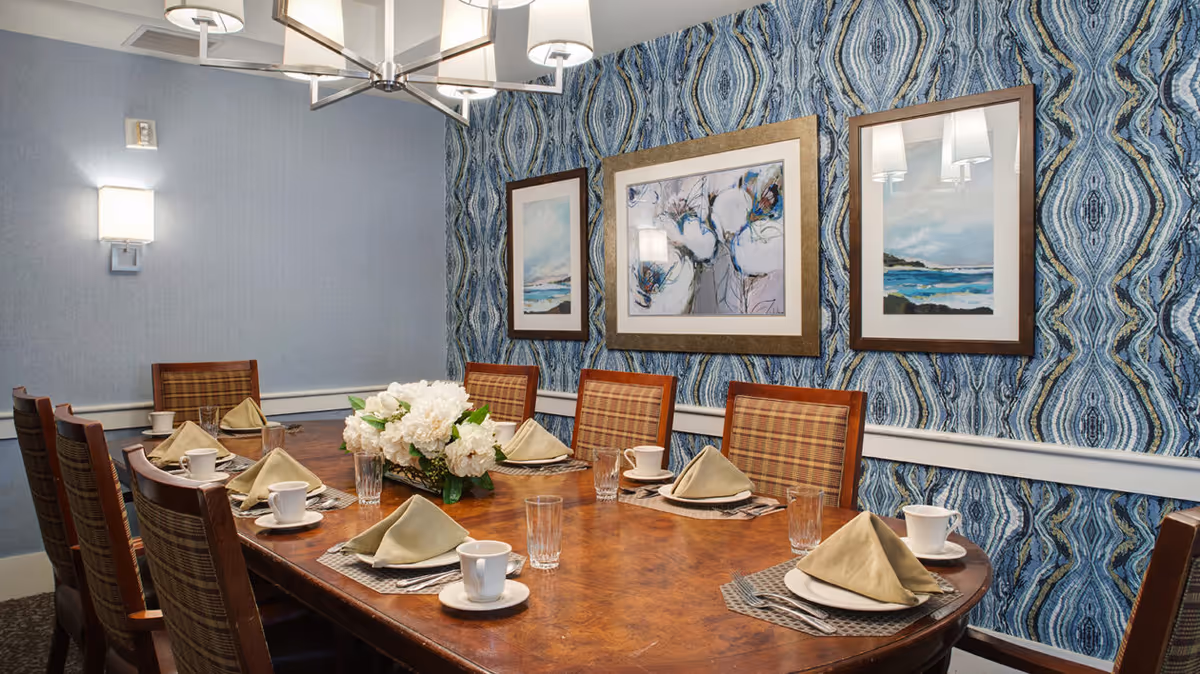 A dining room with a wooden table set for eight people. Each place setting has a beige napkin folded on a white plate, a cup and saucer, a glass, and silverware on a woven placemat. A floral centerpiece with white flowers is in the middle of the table. The walls have blue patterned wallpaper with three framed paintings hanging. A modern chandelier with multiple lights hangs above the table.