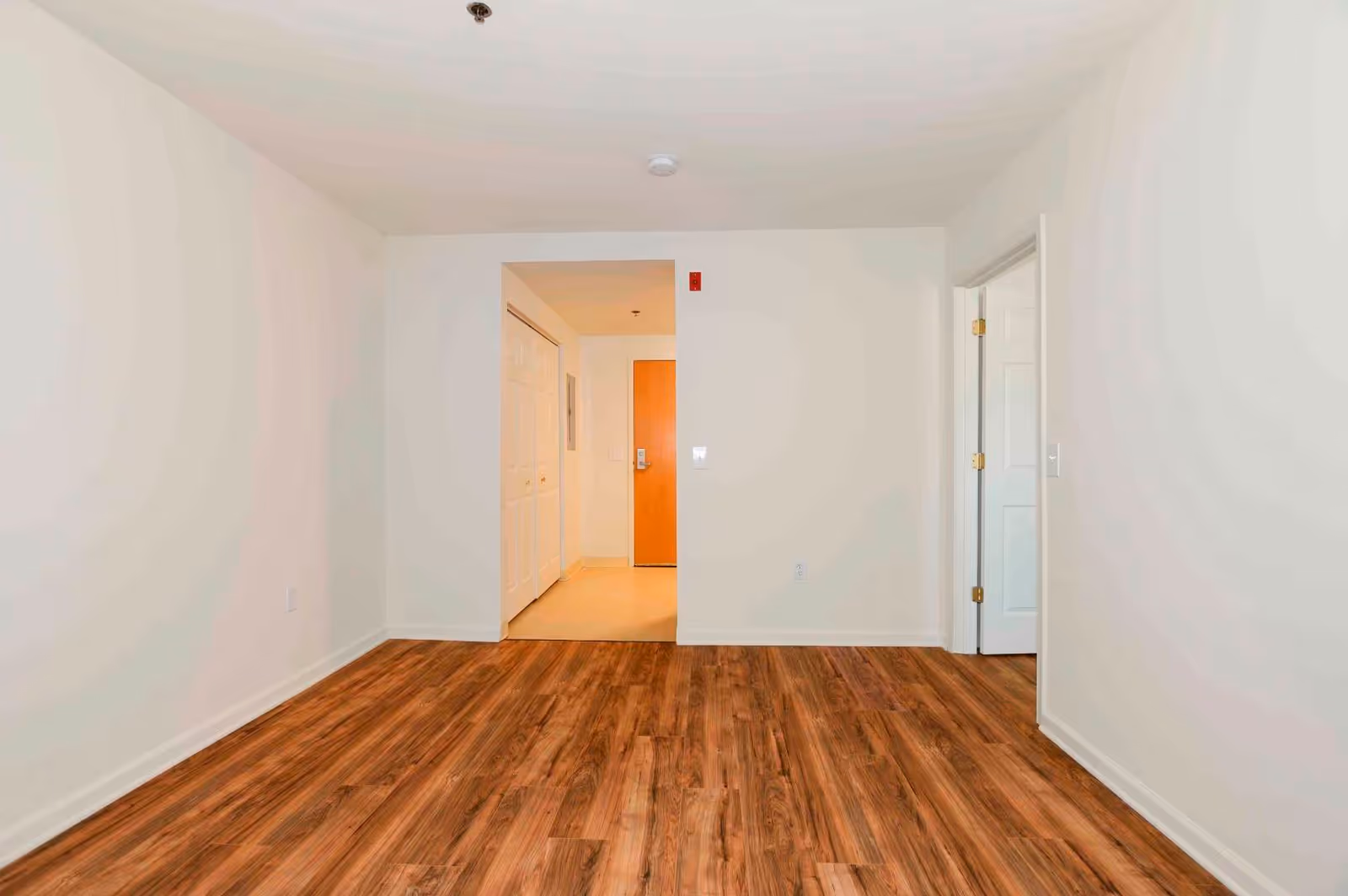 Empty room with wood flooring and white walls, showing a doorway leading to a hallway with a wooden door and closet with white double doors.