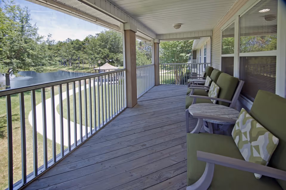 Covered outdoor balcony with wooden floor and railing overlooking a pond and green trees. The balcony has several green cushioned chairs with patterned pillows and small round tables between them.