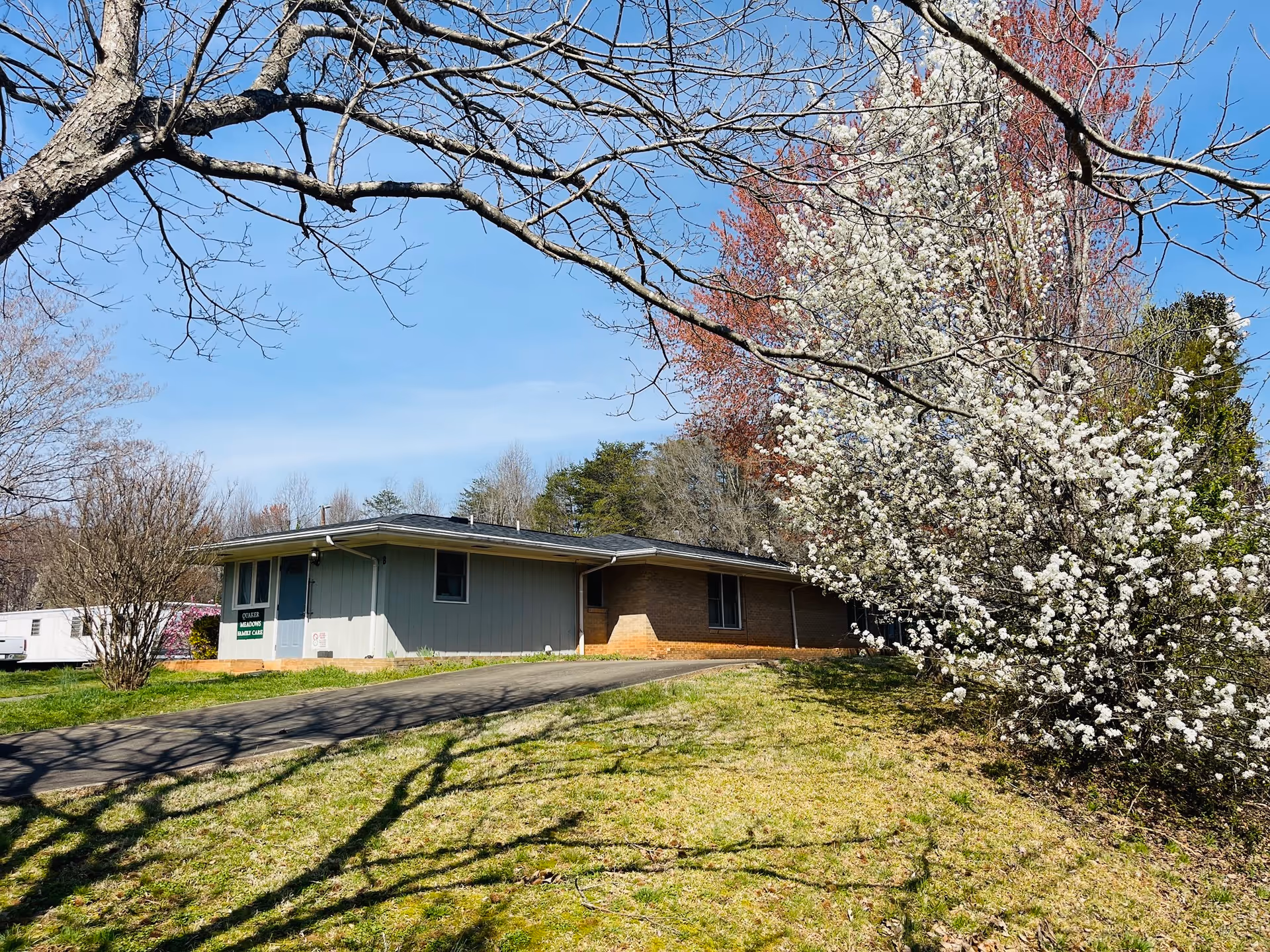A single-story building with a light blue and brick exterior surrounded by trees and bushes, some with white blossoms, under a clear blue sky. A paved driveway leads up to the building, which has a sign that reads 'Quaker Meadows Family Care Home.'