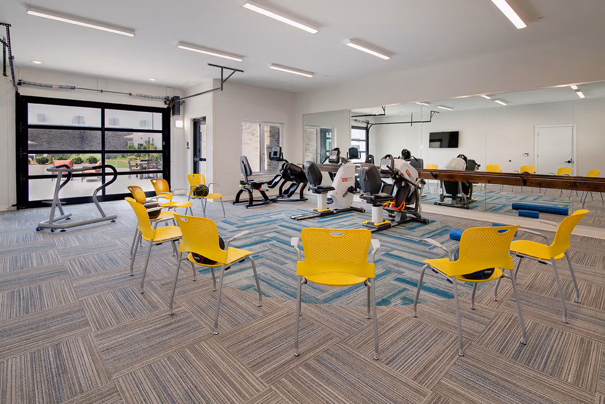 A bright exercise room with yellow chairs arranged in a circle, several recumbent exercise bikes, a treadmill, and a large wall mirror. The room has a patterned carpet, white walls, and large windows allowing natural light to enter. There is a TV mounted on the wall and some exercise mats on the floor.