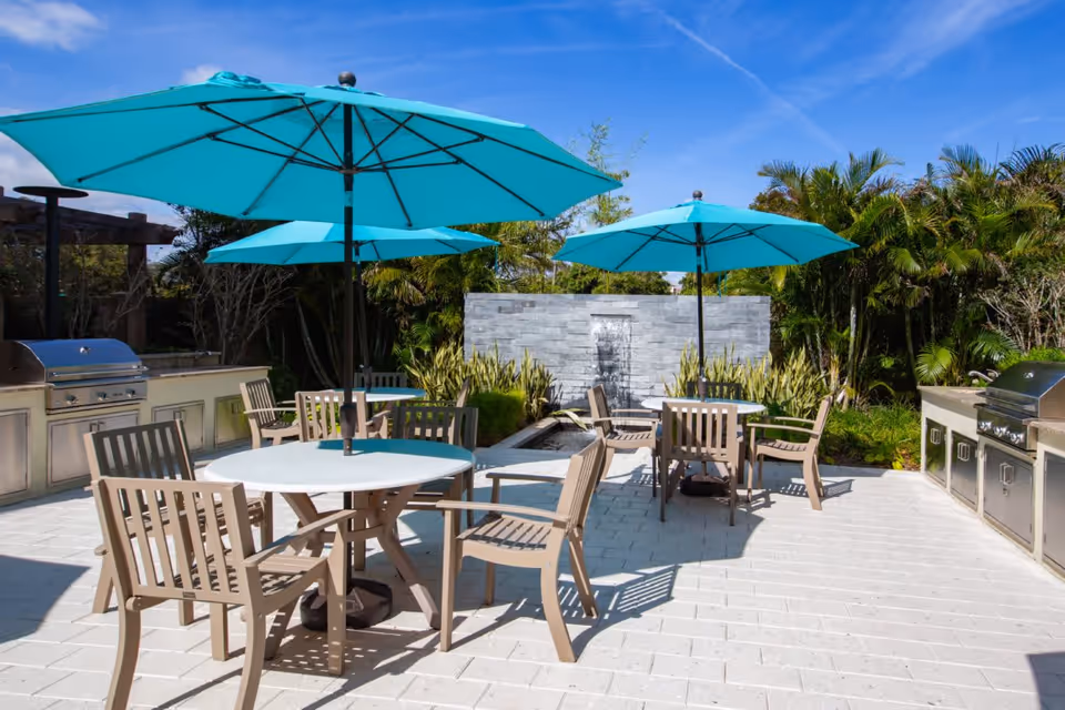 Outdoor patio area with round tables and beige chairs under large turquoise umbrellas. There are built-in stainless steel grills on both sides and a water feature with a stone wall and plants in the background, surrounded by greenery and palm trees.