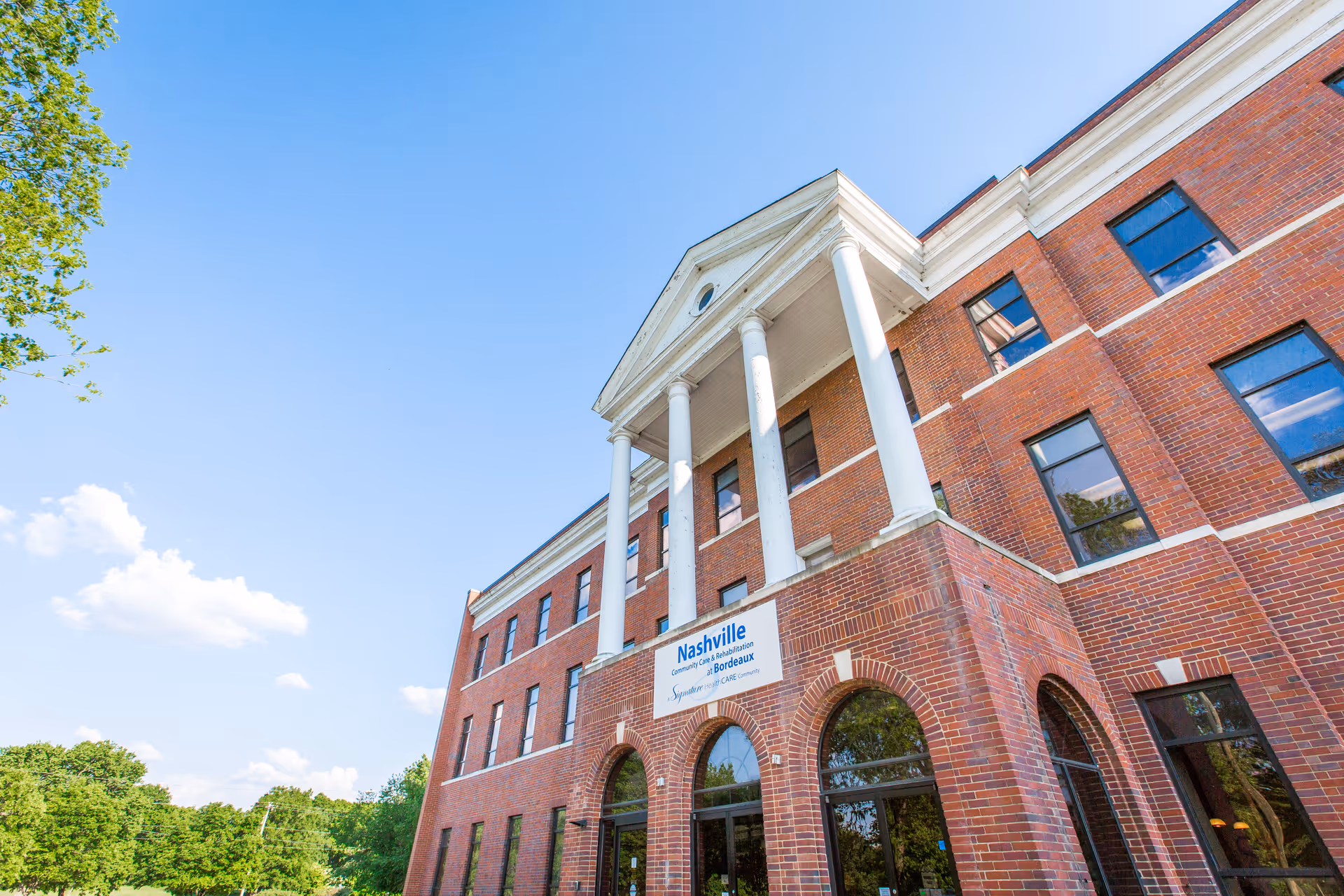 Exterior view of a three-story red brick building with white columns at the entrance under a clear blue sky. The building has multiple windows and a sign that reads 'Nashville Community Care & Rehabilitation at Bordeaux'. Trees and greenery are visible around the building.