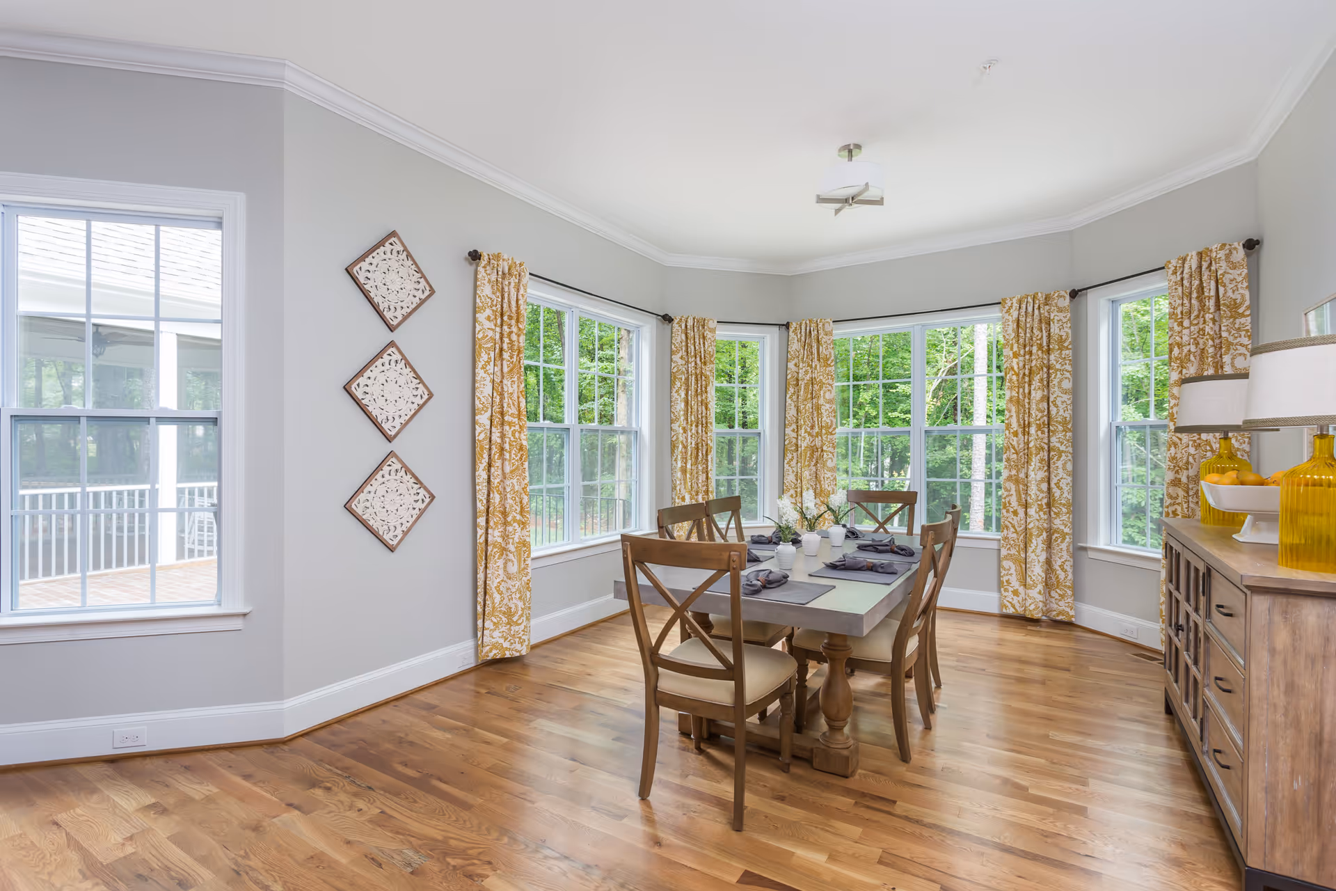 A bright dining room with large windows featuring yellow patterned curtains. The room has a wooden dining table set with six chairs, place settings, and a centerpiece. There is a wooden sideboard with two yellow glass lamps and decorative items. The floor is hardwood, and the walls are painted light gray.