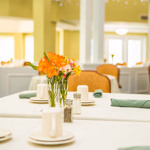 A dining table set with white tablecloth, white cups on saucers, green cloth napkins, salt and pepper shakers, and a vase with orange and yellow flowers in a bright dining room with soft yellow walls and cushioned chairs.