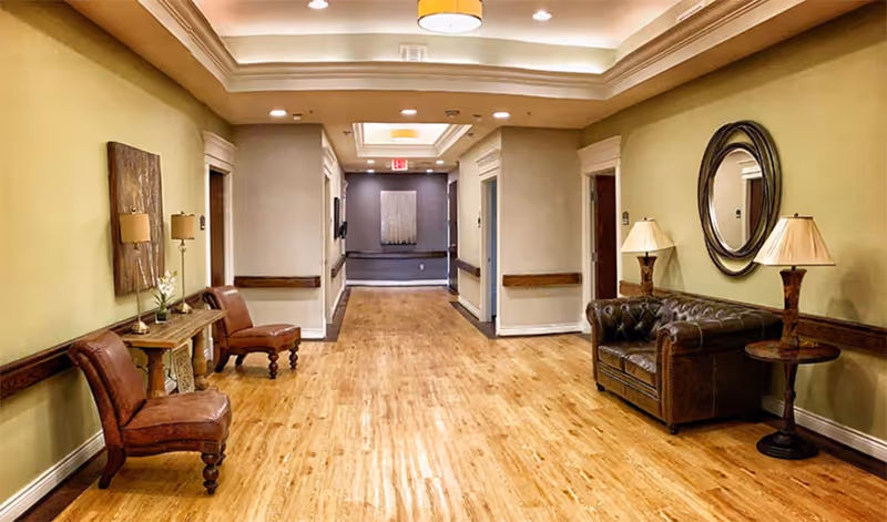 Well-lit interior hallway with wooden floors, leather seating, side tables, lamps and a wall mirror.