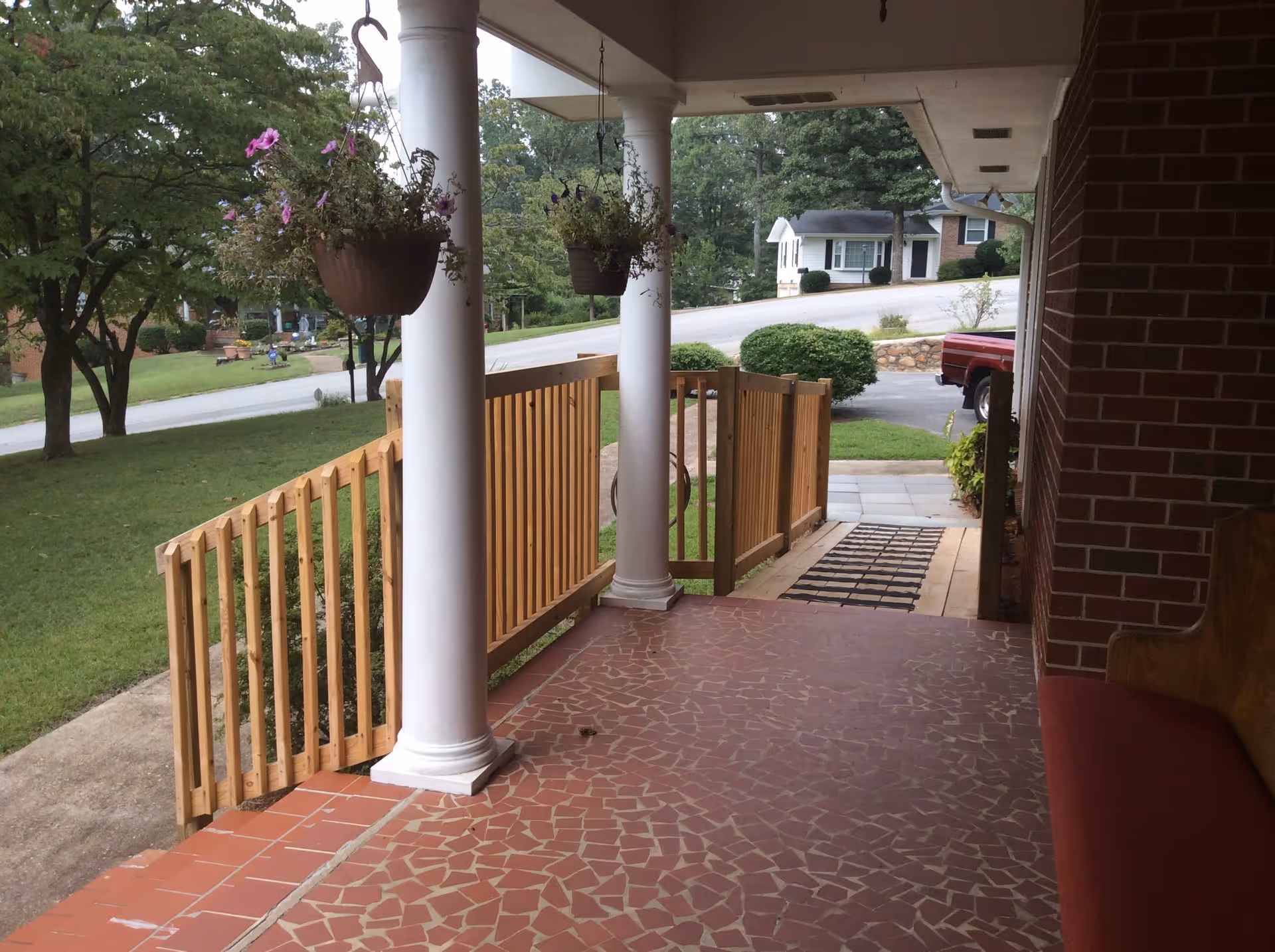 View of a covered porch with mosaic tile flooring, two white columns, hanging flower pots with purple flowers, a wooden railing and gate, and a red cushioned bench against a brick wall. The porch overlooks a green lawn, trees, a street, and houses in the background.