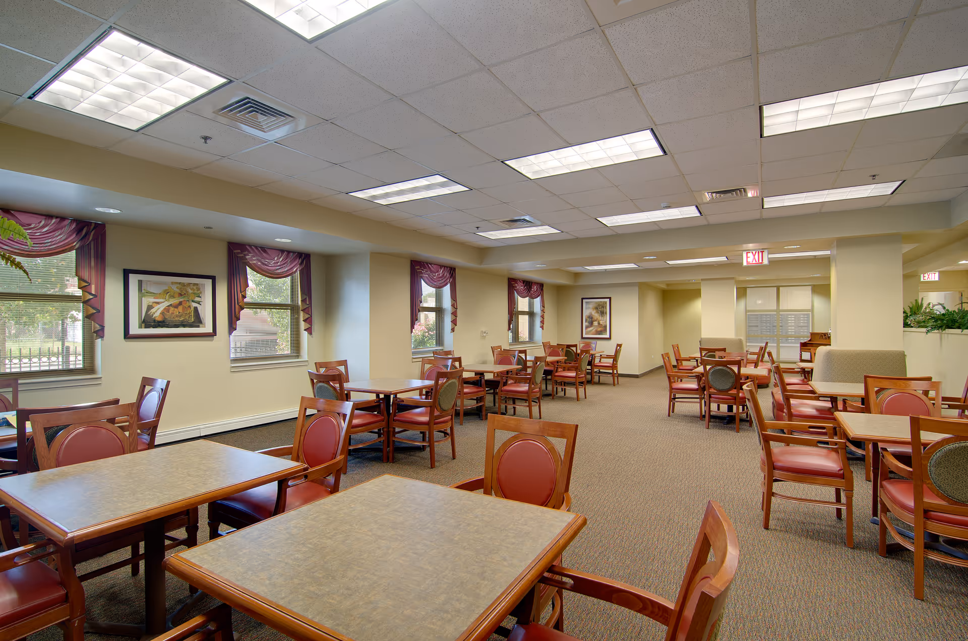 A spacious dining room with multiple wooden tables and chairs featuring red cushioned seats. The room has beige walls, carpeted floors, and several windows with purple valances allowing natural light to enter. Ceiling lights illuminate the area, and framed artwork decorates the walls. Exit signs are visible in the background.