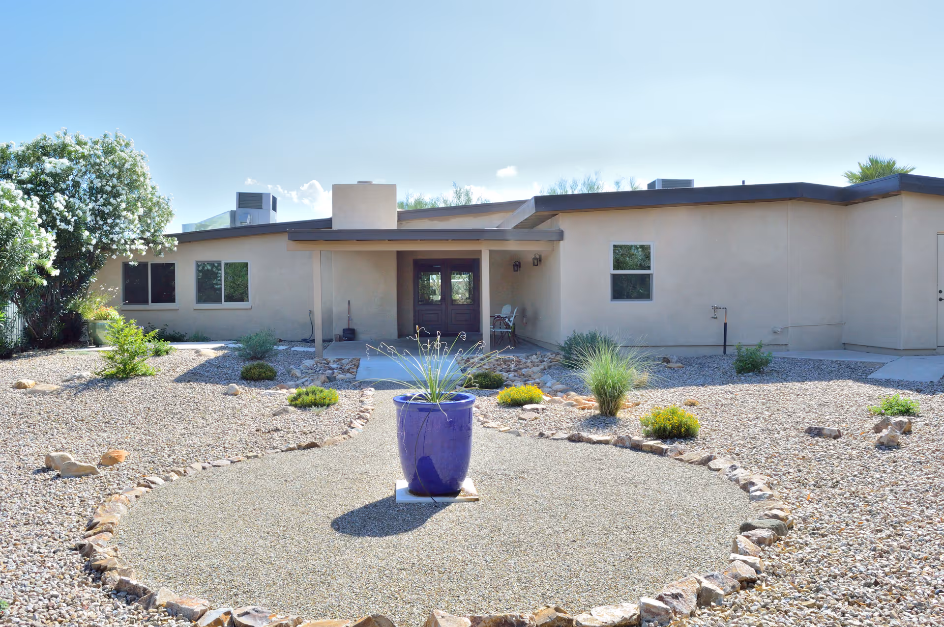 Exterior view of a single-story building with beige walls and a flat roof, surrounded by a landscaped yard with gravel, small bushes, and a large blue planter with a plant in the center of a circular gravel area.