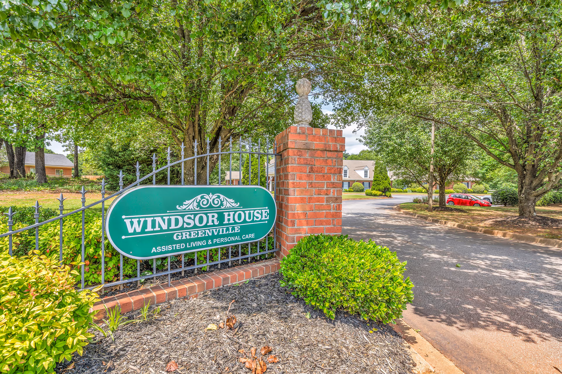 Green Windsor House Greenville entrance sign mounted on a fence and brick pillar beside a tree-lined driveway.