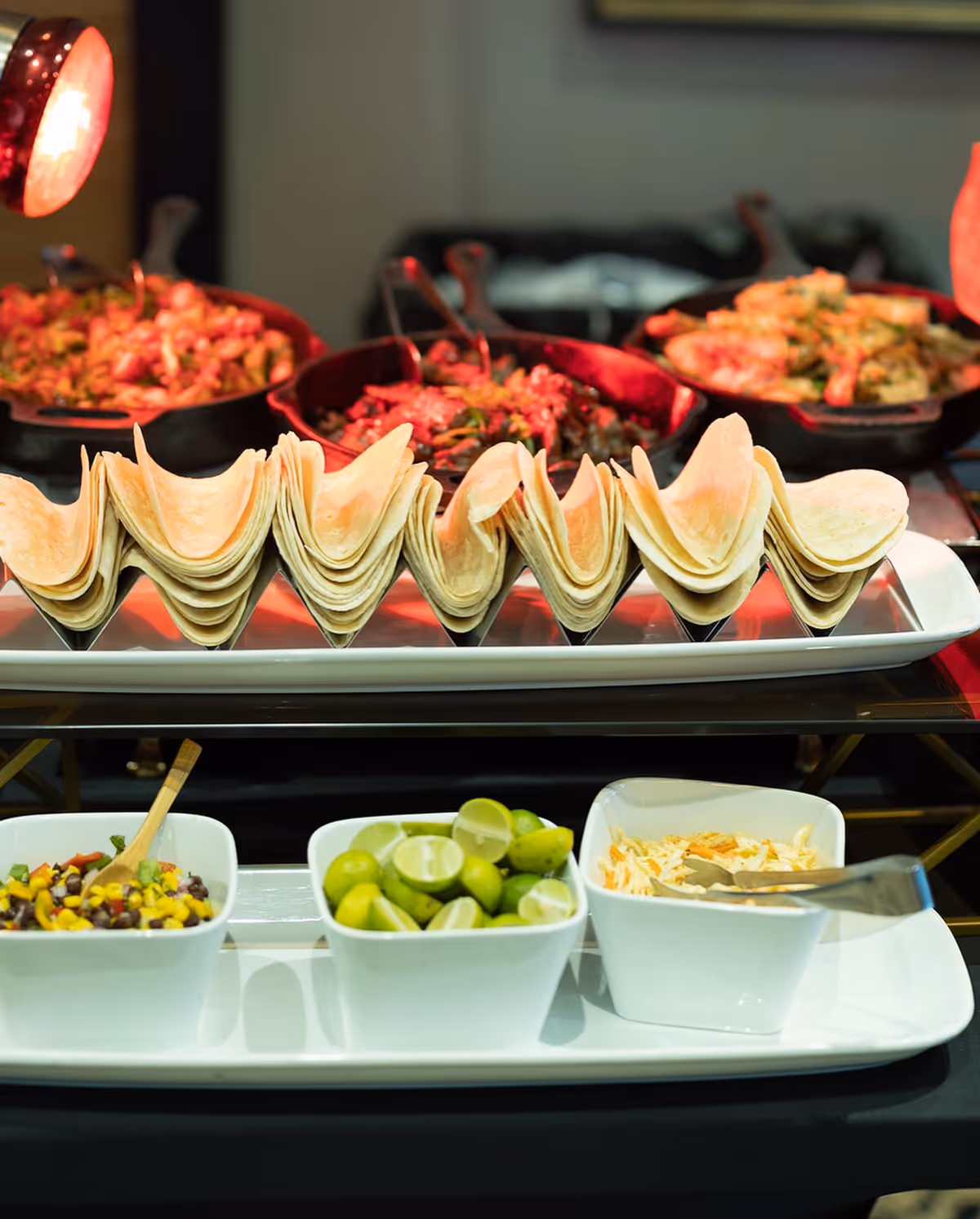 Stacked tortillas arranged on a buffet tray with bowls of lime wedges, salsa, and shredded cheese and heated pans of food in the background.