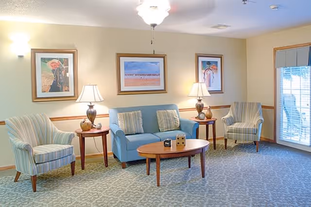 A cozy living room with a light blue sofa and two striped armchairs arranged around a wooden coffee table. Two side tables with lamps flank the sofa. Three framed pictures hang on the beige walls, and a window with blinds lets in natural light.