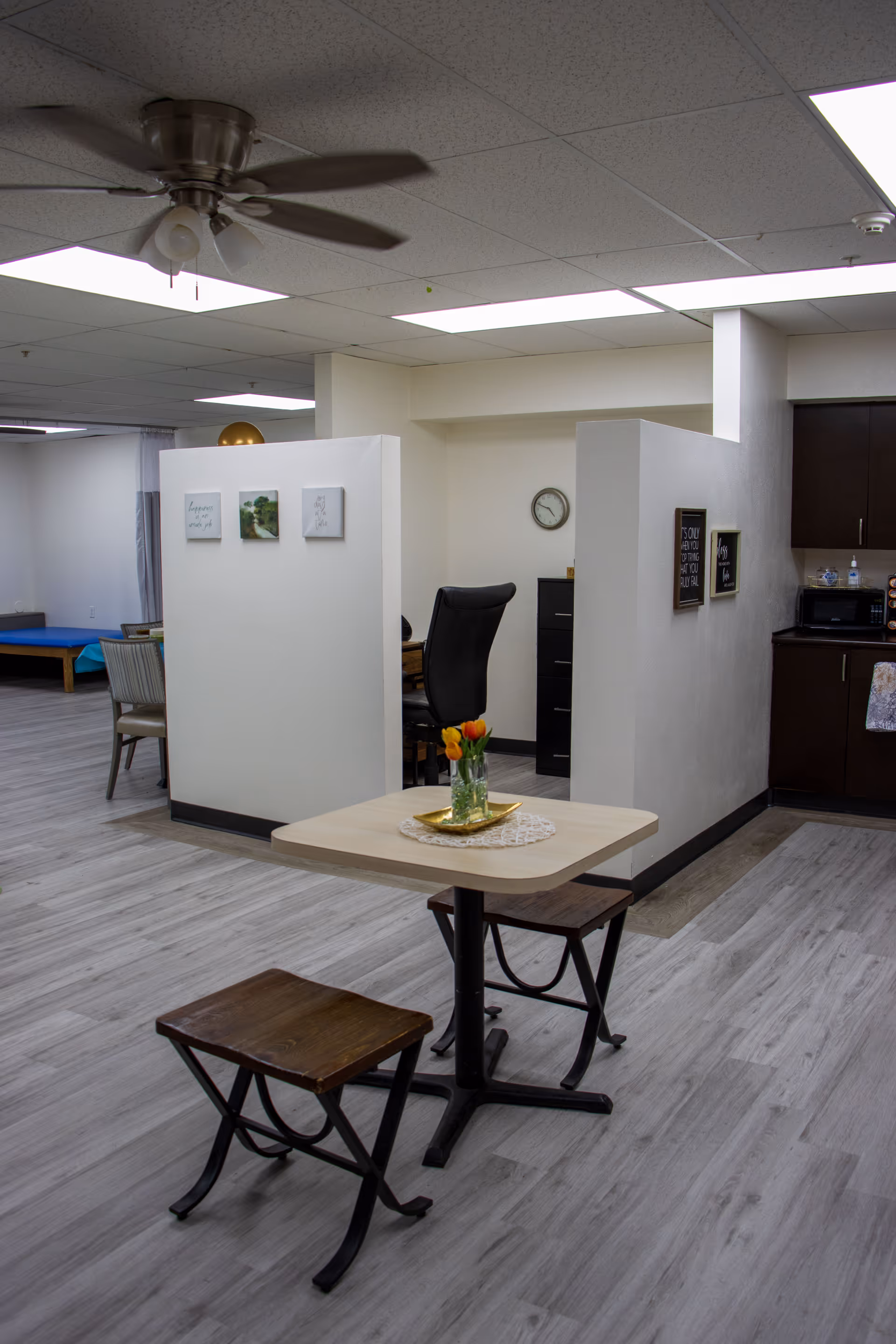 Interior view of a senior living facility showing a small square table with a vase of orange tulips on a doily, two wooden stools, a ceiling fan, and partial views of a kitchenette and office area with a black office chair and filing cabinet.