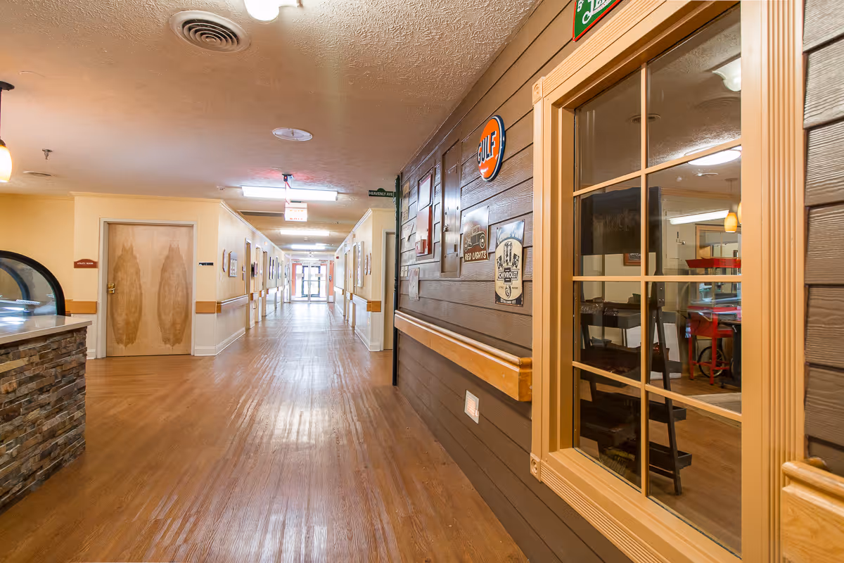 A long, well-lit hallway in a senior living facility with wooden floors and beige walls. On the right side, there is a brown wooden wall decorated with vintage signs and a window showing a room with chairs and tables. The left side has a counter with a stone facade and wooden doors along the hallway. The hallway extends to a glass exit door at the far end.