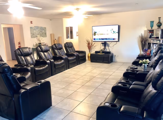 A large communal TV room with two rows of black leather recliners facing a wall-mounted television.