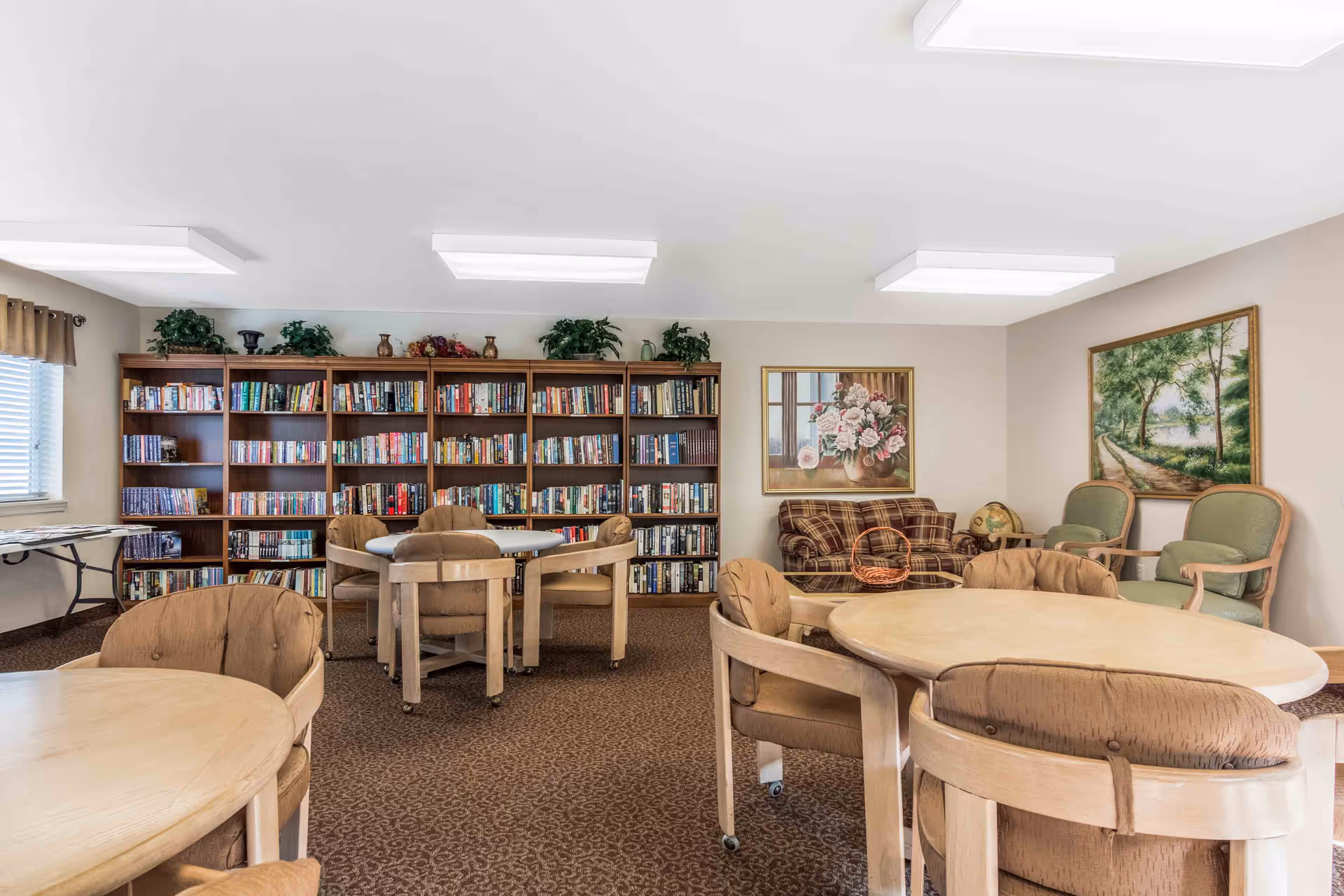 A cozy common room with round tables and cushioned chairs on wheels, a large bookshelf filled with books along the back wall, a plaid sofa, two green armchairs, and framed paintings of flowers and a nature path on the walls.