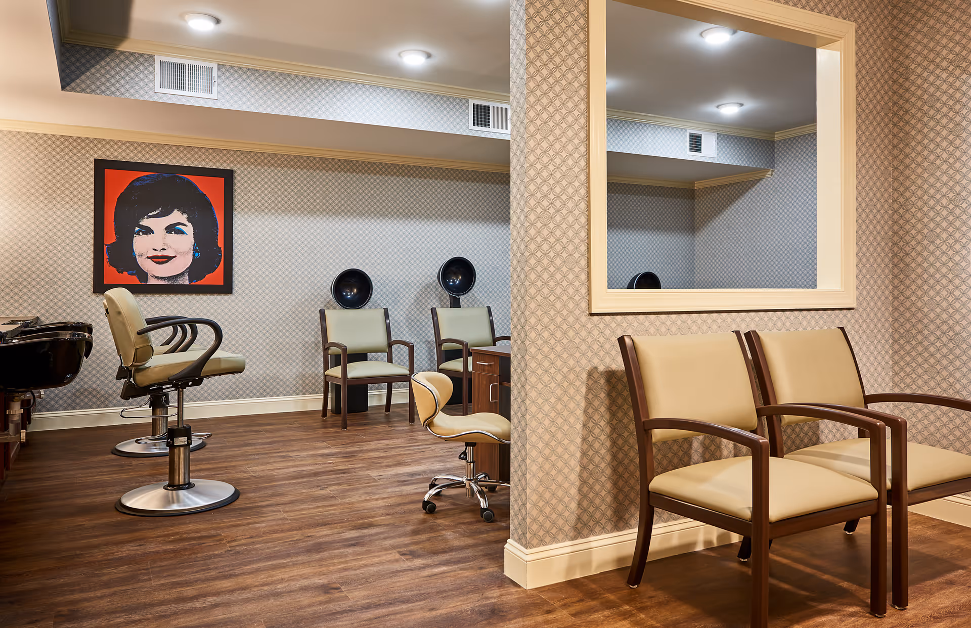 Interior view of a salon area in a senior living facility with beige and brown chairs, a large mirror on the wall, two hair drying stations, and a framed pop art portrait of a woman with dark hair and red background. The floor is wooden and the walls have patterned wallpaper.