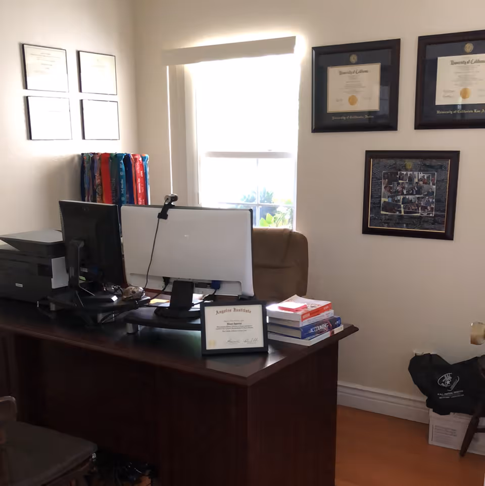 An office space with a wooden desk holding two computer monitors, a printer, a framed certificate, and a stack of books. Behind the desk is a brown office chair, and the wall features framed diplomas and a collage of photos. A window with a white blind is centered on the wall.