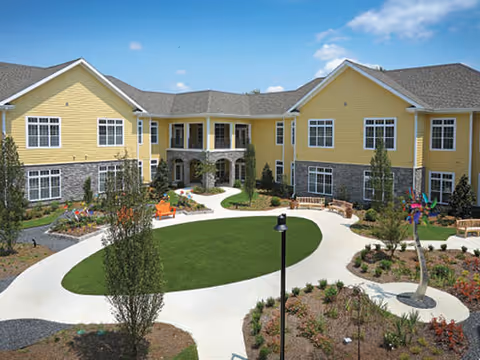 Landscaped sunlit courtyard with a circular walkway and seating, surrounded by a two-story yellow residential building.