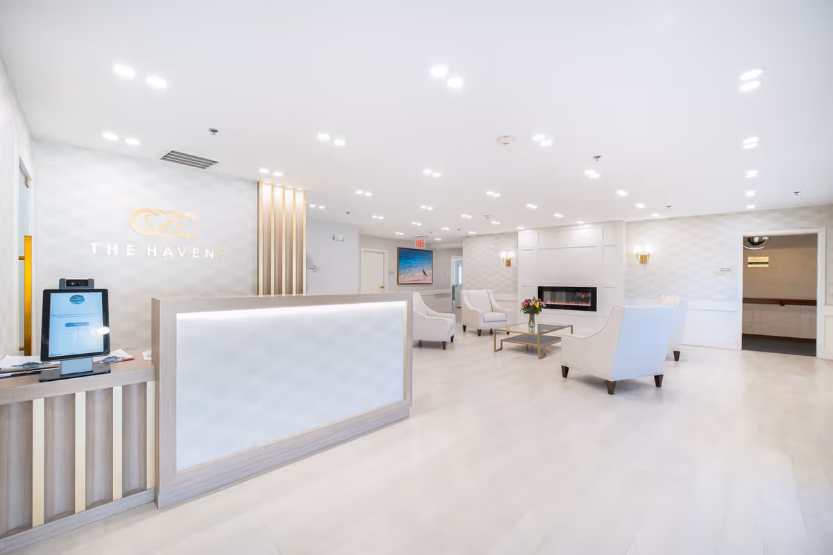 Bright and spacious reception area of The Havens Assisted Living with a modern front desk, white armchairs arranged around a glass coffee table with a flower vase, a wall-mounted electric fireplace, and soft lighting fixtures on the walls and ceiling.