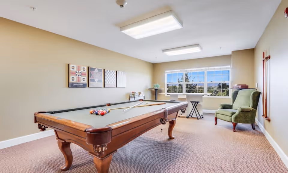 A well-lit game room with a pool table in the center, a green armchair to the right, a small table with a plant near a large window showing trees outside, and framed artwork on the beige walls.