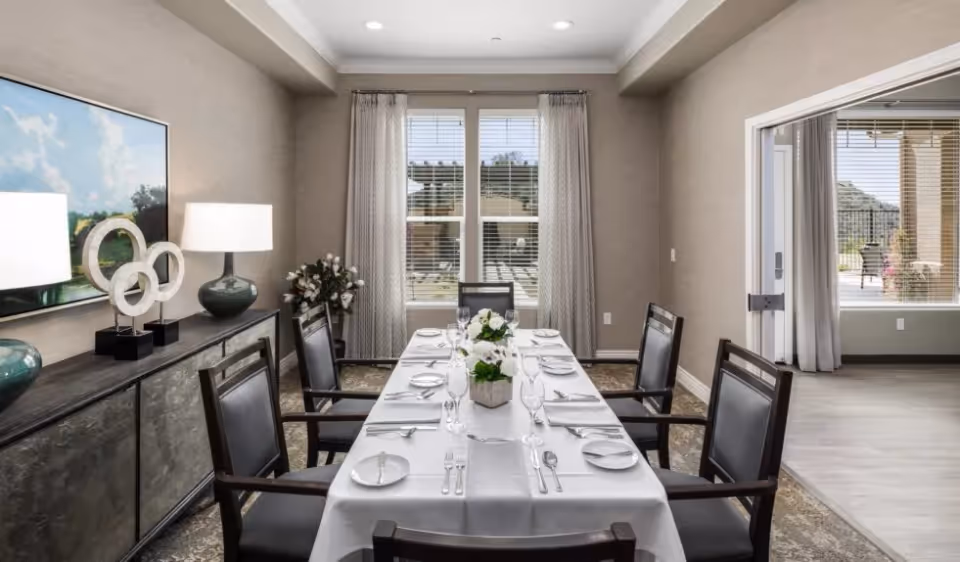 A formal dining room with a long table set for a meal, surrounded by chairs, a sideboard with lamps and decorative items, and windows with curtains.