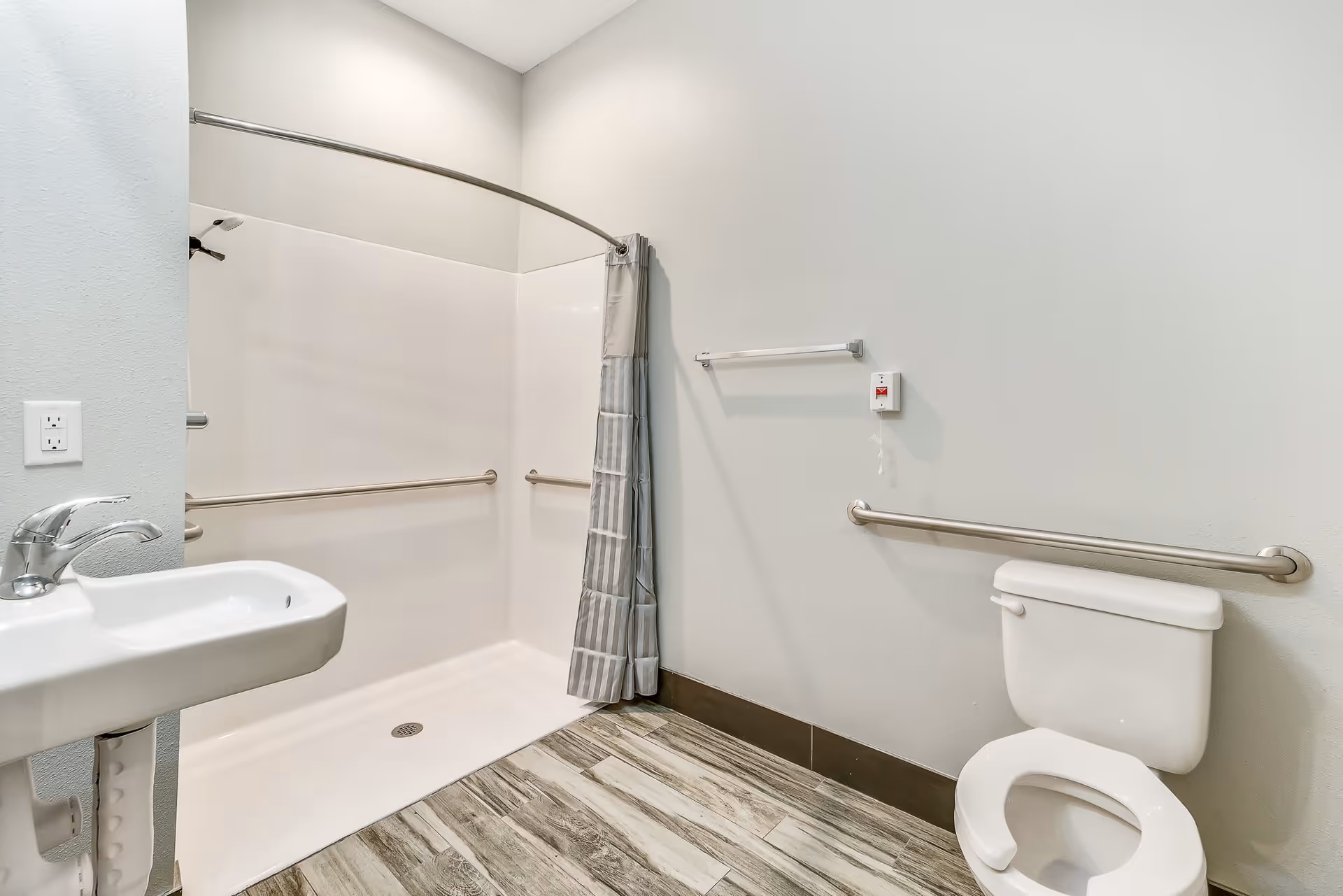 A clean, accessible bathroom with a white toilet, a wall-mounted sink, and a walk-in shower with grab bars and a curved shower curtain rod. The floor has wood-like tiles, and the walls are painted light gray.