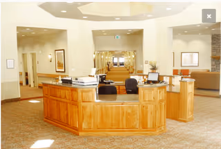 Reception area with a wooden front desk featuring office equipment and chairs. The space has beige walls, carpeted floors, and recessed lighting. Hallways and seating areas are visible in the background.