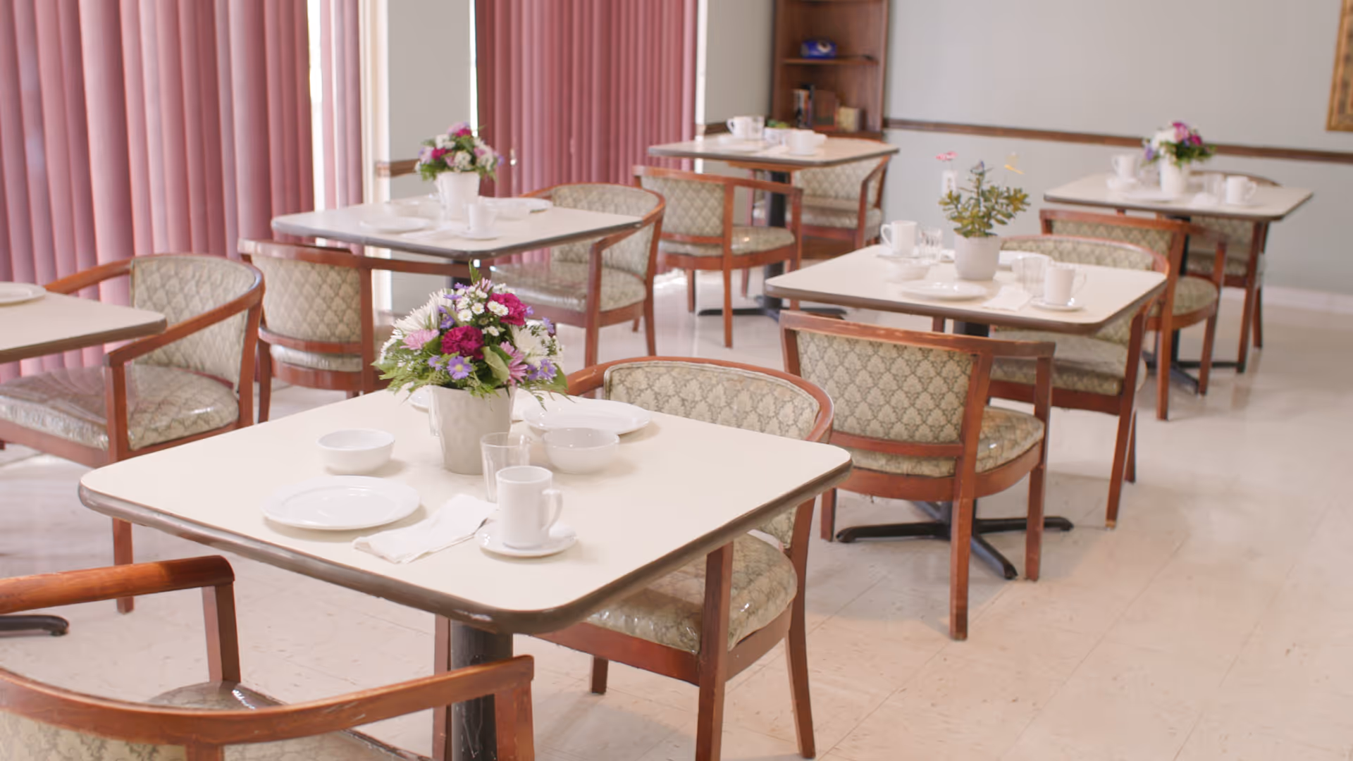 A dining area with several square tables, each set with white plates, cups, and napkins. Each table has a small floral centerpiece. The chairs have wooden frames with patterned upholstery. Pink vertical blinds cover the windows in the background.