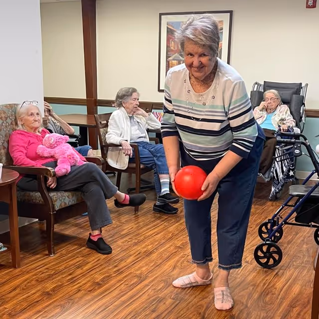 A group of elderly residents in a senior living common room watch as a woman bends forward holding a red ball, surrounded by chairs, walkers, and wood flooring.